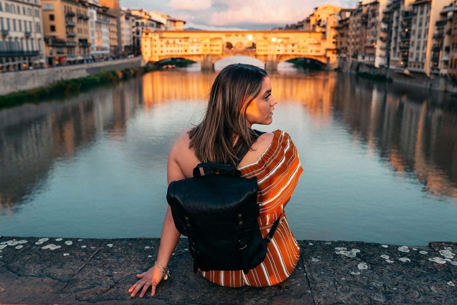 A traveler sits on a stone embankment above a river at golden hour.