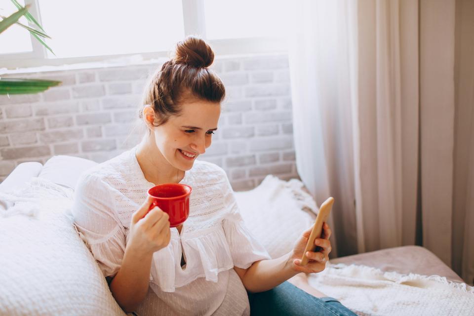 A woman rests in bed with tea and her phone.